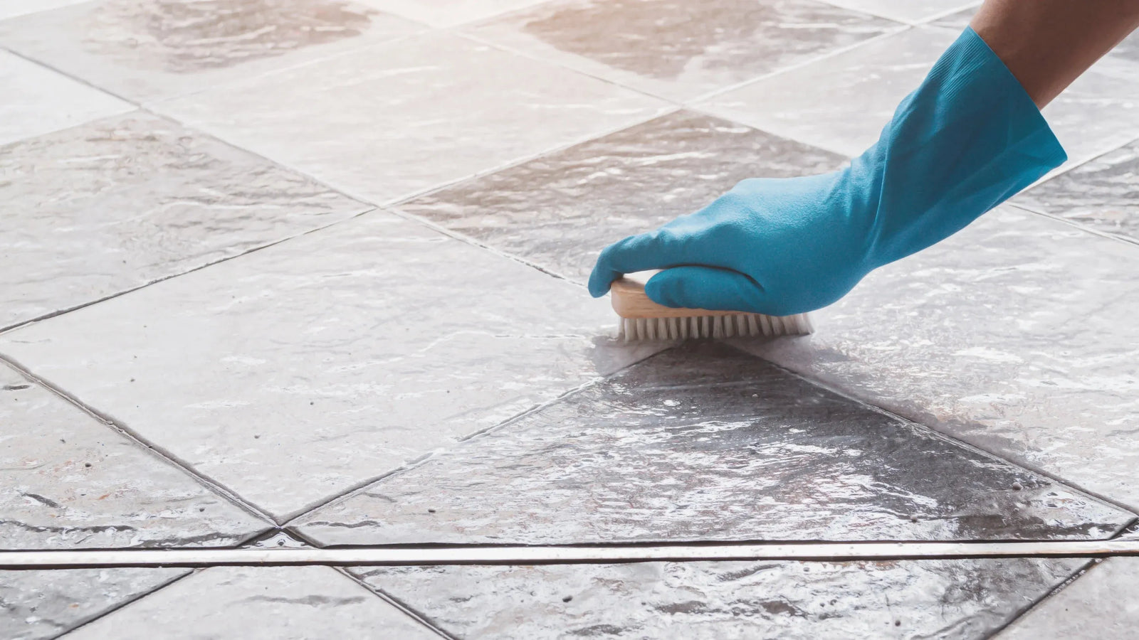 Person wearing a blue glove scrubbing tile grout with a brush.
